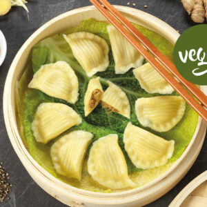 Ein Bambusdämpfer mit veganen DIM SUM Red Curry-Knödeln auf Kohlblättern, daneben Essstäbchen, Sojasauce, Ingwer, Pfeffer und Bohnensprossen. Rechts erscheint ein grünes veganes Etikett.