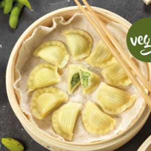 Draufsicht auf DIM SUM Red Curry in einem Bambusdämpfer mit veganen Teigtaschen, von denen eine aufgeschnitten ist, um die grüne Füllung freizulegen. Essstäbchen liegen auf dem Dampfgarer. In der Nähe befinden sich Tofuwürfel, Sojasauce, Edamame und ein grünes "Vegan"-Etikett auf einer Schiefertafel.