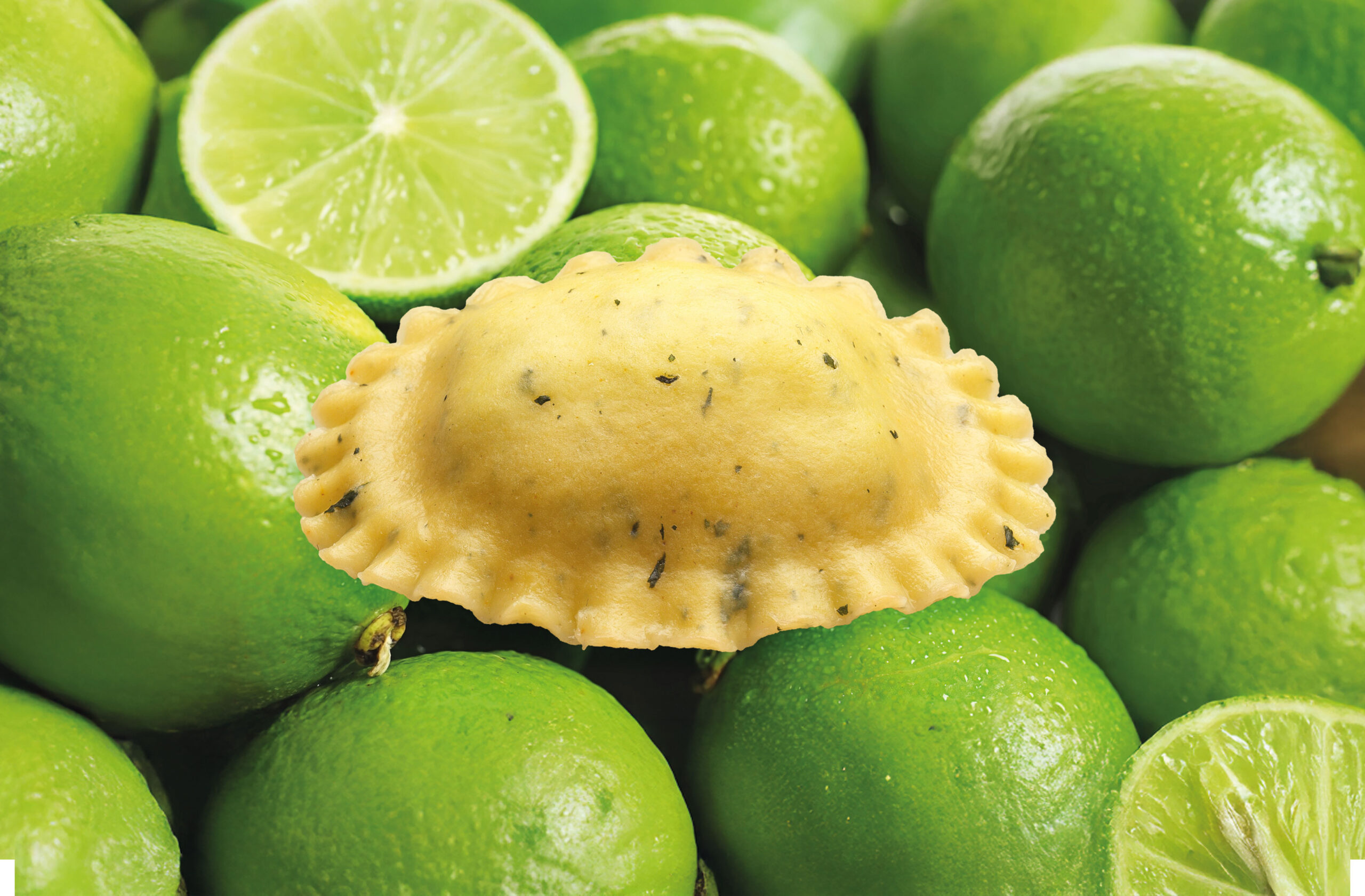 In der Mitte befindet sich ein einzelnes St&uuml;ck Ravioli-Nudeln, umgeben von ganzen und in Scheiben geschnittenen gr&uuml;nen Limetten im Hintergrund. Die Ravioli sind scharf abgebildet, w&auml;hrend die Limetten leicht verschwommen sind.