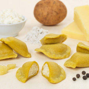 Ein Arrangement aus Bergkäse-Schlutzern - ungekochte Ravioli mit Käse- und Kartoffelfüllung -, umgeben von Käsestücken, schwarzen Pfefferkörnern, gewürfelten und ganzen Kartoffeln, Mehl und einer Schüssel mit Ricotta-Käse.