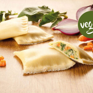 Drei Vegane Maultaschen, eine davon aufgeschnitten, um die Gemüsefüllung freizulegen, umgeben von gewürfelten Karotten, Spinatblättern, Karottenspitzen und roten Zwiebeln auf einer Holzunterlage. Auf der rechten Seite erscheint ein grünes Etikett mit der Aufschrift "vegan".