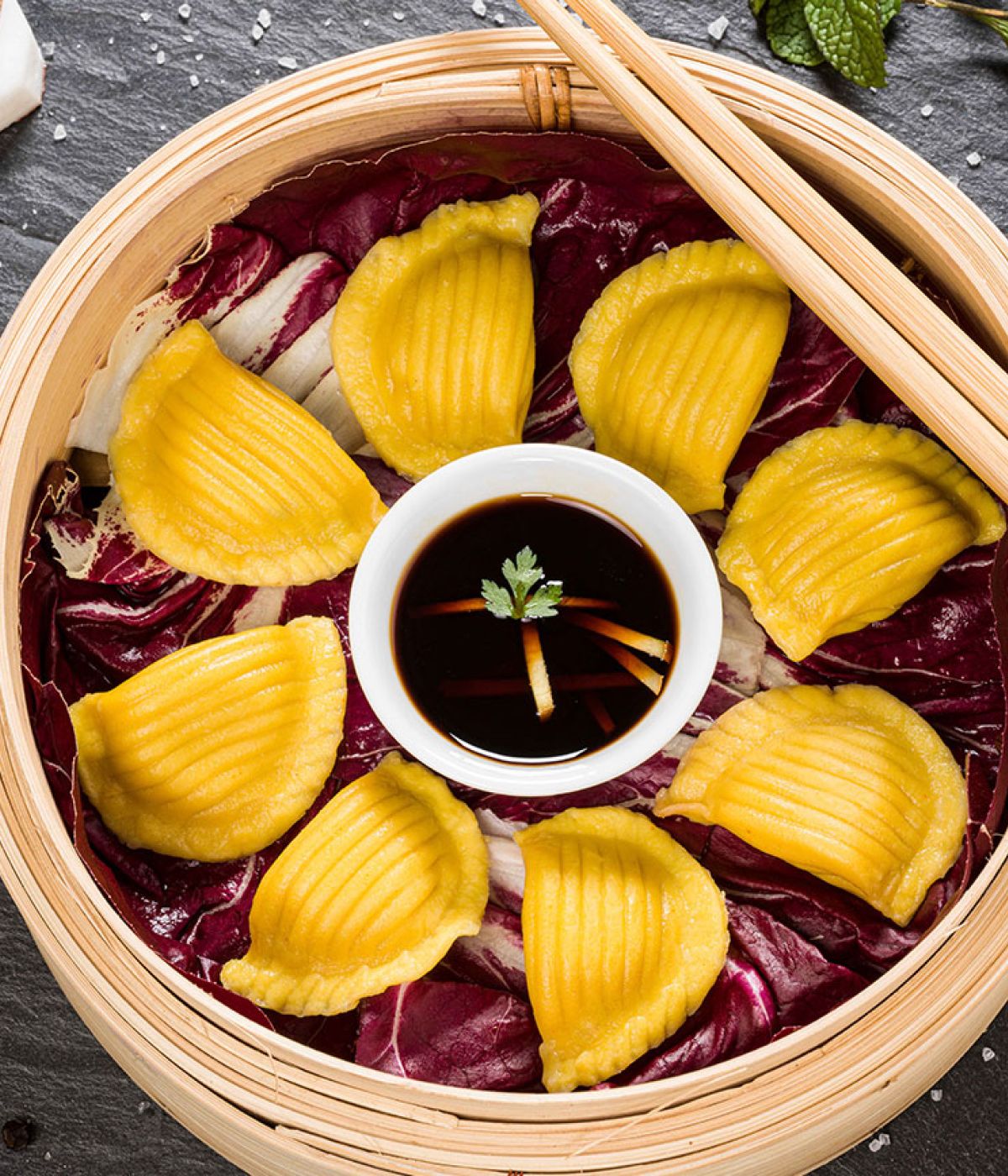 Ein Bambusd&auml;mpfer, gef&uuml;llt mit gelben Teigtaschen, die kreisf&ouml;rmig um eine Schale mit Sojasauce angeordnet sind.